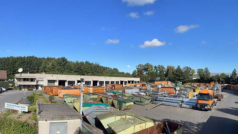 Blick über das moderne Aluminium Krüger Betriebsgelände mit Containern, Aluminiumteilen und einem Firmen-LKW unter blauem Himmel