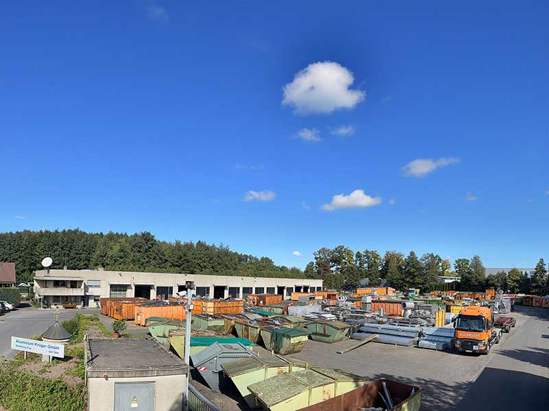 Blick über das moderne Aluminium Krüger Betriebsgelände mit Containern, Aluminiumteilen und einem Firmen-LKW unter blauem Himmel
