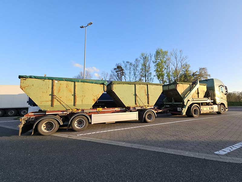 Grüner LKW von Aluminium Krüger mit beladenem Anhänger und mehreren Containermulden auf einem Parkplatz bei Tageslicht